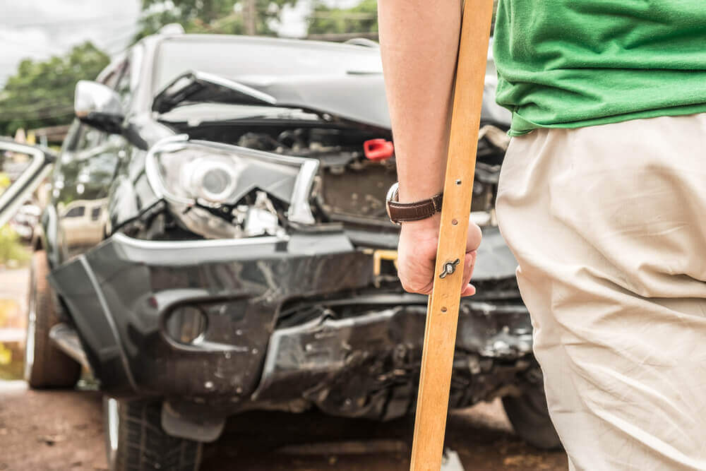 Injured driver on crutches looking into the damaged car from accident.